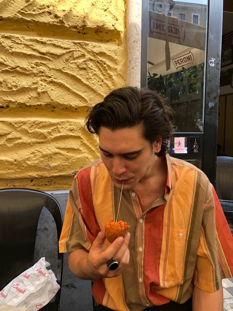 A man sitting outside a restaurant with yellow walls wearing a colourful, striped shirt. He is looking down at the arancini he is holding, that he has taken a bite from and there is a string of cheese going from the ball to his mouth